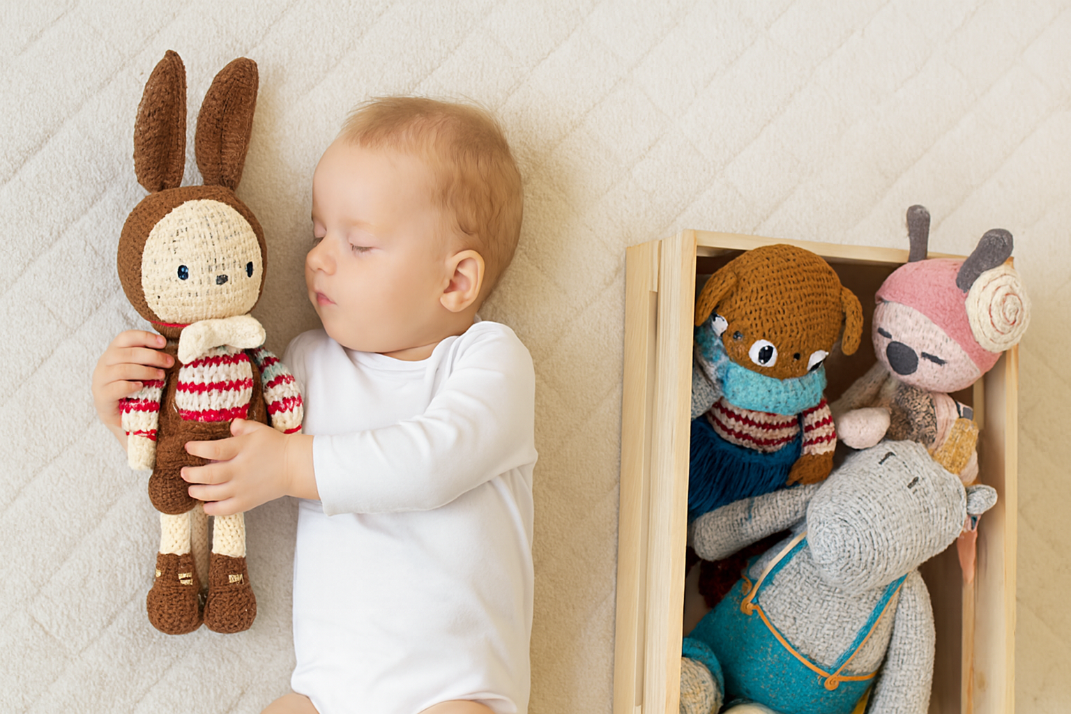 Bébé tenant un doudou au crochet, entouré d'autres peluches artisanales dans une caisse