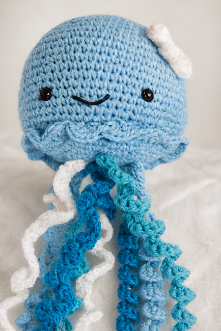 Peluche pieuvre crochetée en coton bleu avec tentacules en spirale bleu foncé, turquoise et blanc, visage souriant et nœud blanc décoratif, sur fond doux