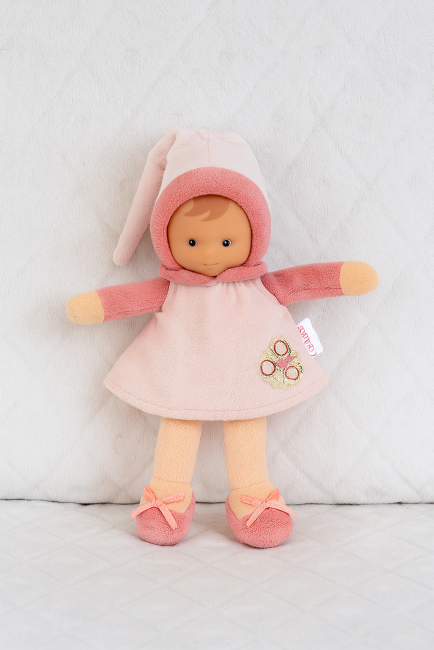 Poupée Corolle en tissu rose avec capuche et broderie florale, posée sur fond matelassé 