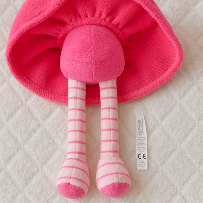 Poupée en peluche rayée – seconde main lavée – 27 cm – conforme CE Poupée en peluche rose avec jambes rayées, allongée sur couverture blanche, étiquette CE visible