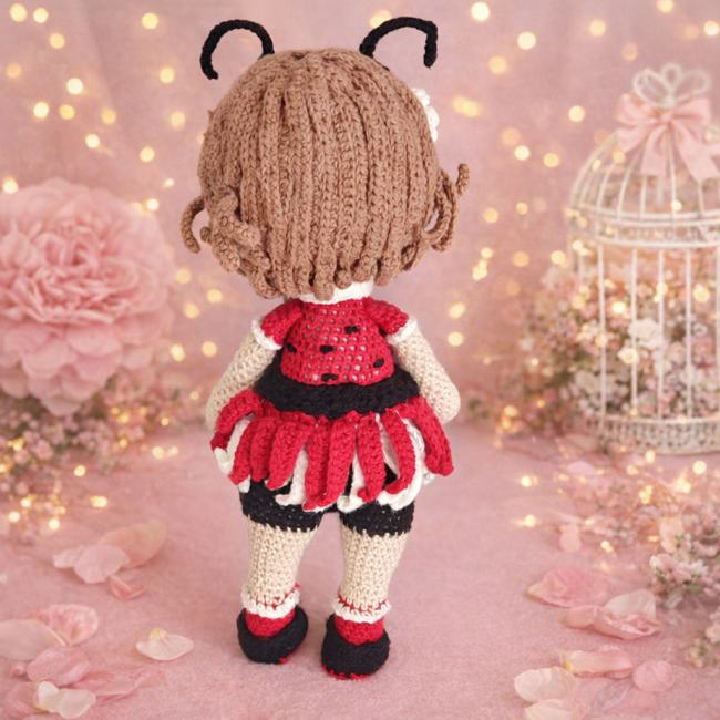 Poupée coccinelle en crochet vue de dos, tenue rouge et noire, décor fleuri rose et guirlande lumineuse.