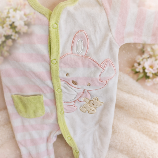 Pyjama naissance rayé rose et blanc avec broderie animale, posé près d’une peluche lapin sur surface douce