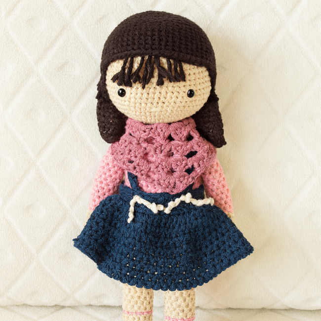 Poupée crochetée artisanale en jupe bleue et haut rose, sur fond blanc texturé