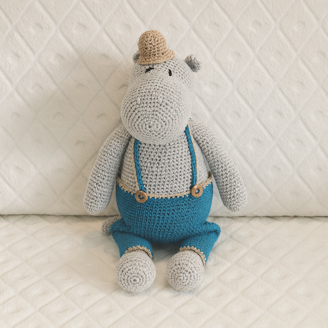 Doudou crocheté, tenue bleue à bretelles croisées, assis sur couverture blanche à motif losange