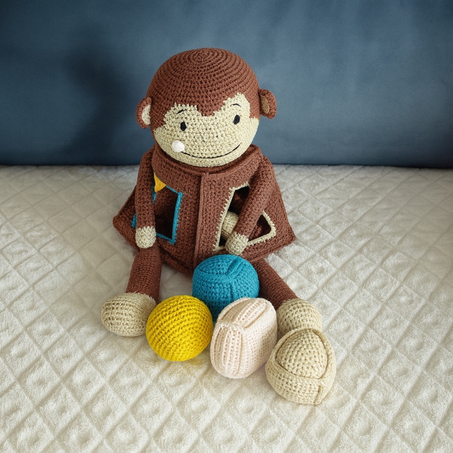 Doudou singe crocheté brun et beige, assis avec quatre balles crochetées colorées, présenté dans une mise en scène douce et lumineuse.