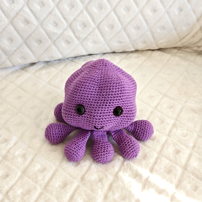 Jouet pieuvre crocheté violet avec yeux noirs et sourire, posé sur couverture blanche
