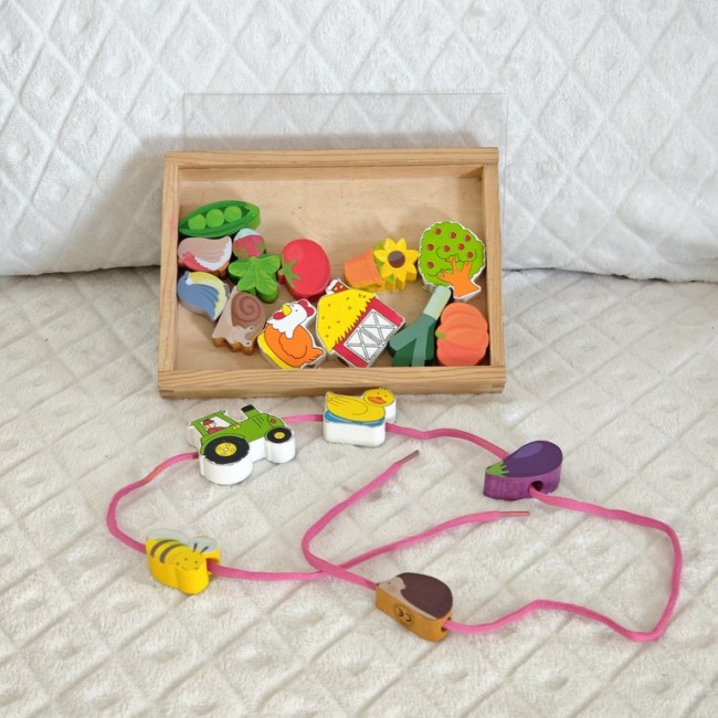Coffret en bois de perles éducatives sur le thème de la ferme, avec pièces colorées enfilées sur un cordon rose, présenté sur fond quilté blanc.