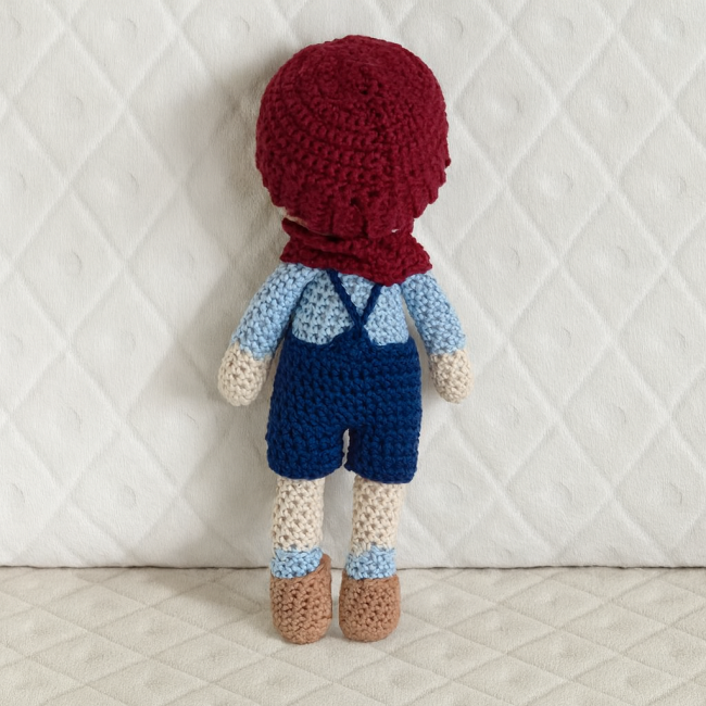 Vue arrière de la poupée crochetée et ses vêtements amovibles Dos de la poupée crochetée avec bonnet bordeaux, salopette bleu marine et chaussures marron