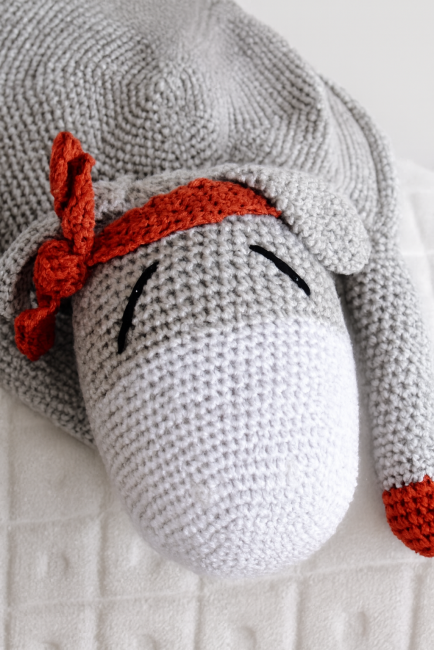 Doudou crocheté représentant un âne gris avec museau blanc, yeux fermés brodés, bandana rouille et oreille rouille.