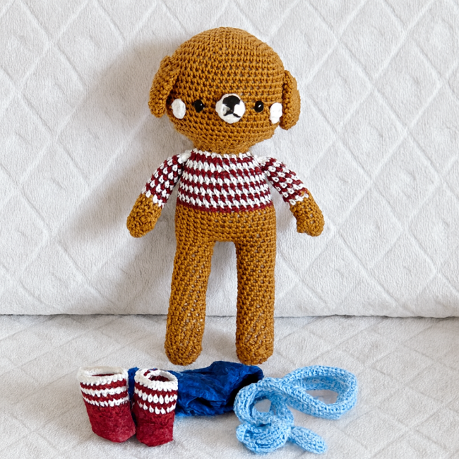Doudou chien crocheté fait main avec vêtements amovibles posés devant sur fond blanc texturé