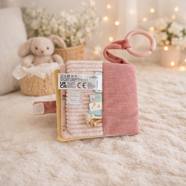 Livre d’éveil sensoriel pastel avec textures, rubans et anneau rose, posé sur surface crème