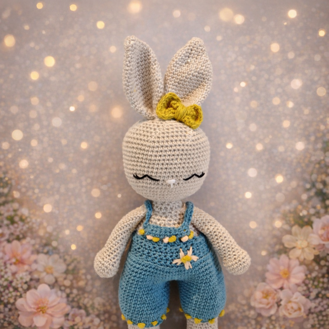 Doudou lapin fait main en crochet, portant une salopette bleue et un nœud moutarde, photographié dans une ambiance chaleureuse avec guirlande lumineuse et fleurs
