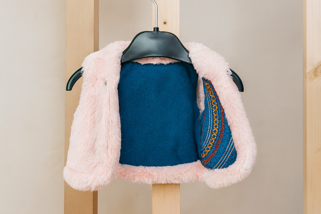 Gilet bébé rose avec intérieur bleu brodé, suspendu sur fond bois clair