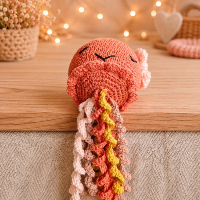Pieuvre en crochet orange aux yeux fermés, ornée d’un noeud , avec tentacules bouclés multicolores et décor flou végétal et lumineux