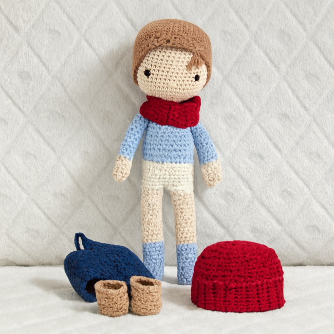 Poupée crochetée avec accessoires amovibles présentés sur fond clair Poupée crochetée garçon avec snood bordeaux et accessoires posés : bonnet, chaussures, sac à dos