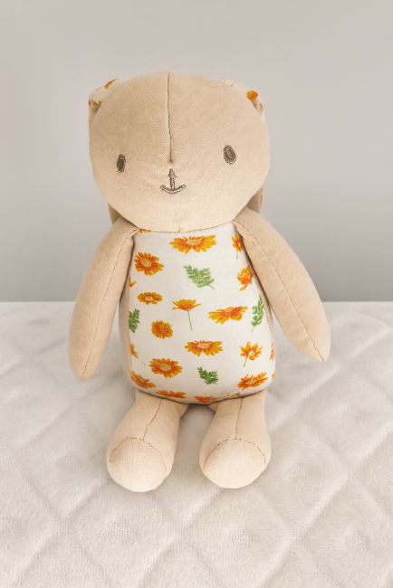 Doudou lapin beige avec ventre et oreilles fleuries, assis sur couverture blanche