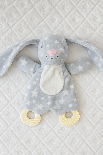 Doudou lapin gris étoilé avec anneaux de dentition crème, posé sur couverture matelassée