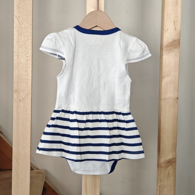 Dos de la robe bébé rayée blanc et bleu avec body intégré Dos d’une robe bébé blanche et bleu marine à rayures avec body intégré, manches courtes, suspendue sur cintre en bois