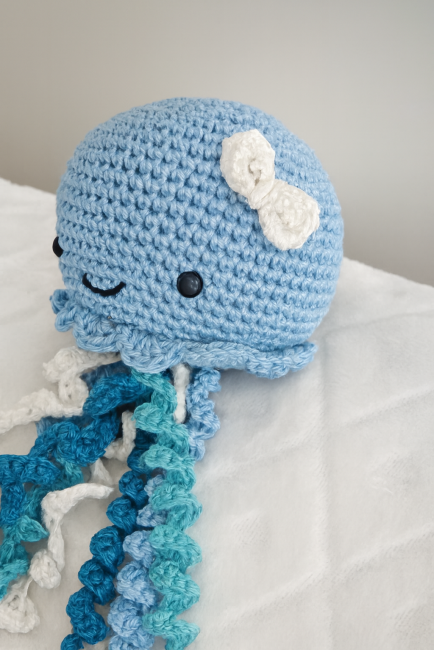 Doudou pieuvre crocheté à la main en coton bleu avec tentacules bouclés bleu, blanc et turquoise, yeux sécurisés et petit nœud blanc, posé sur fond blanc