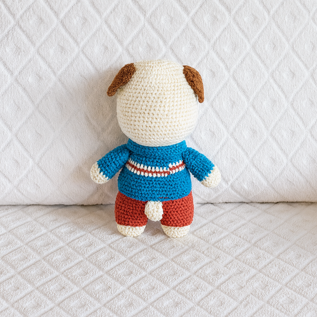 Dos du doudou chien crocheté avec pull bleu rayé et pantalon rouge, sur fond blanc à motif losange