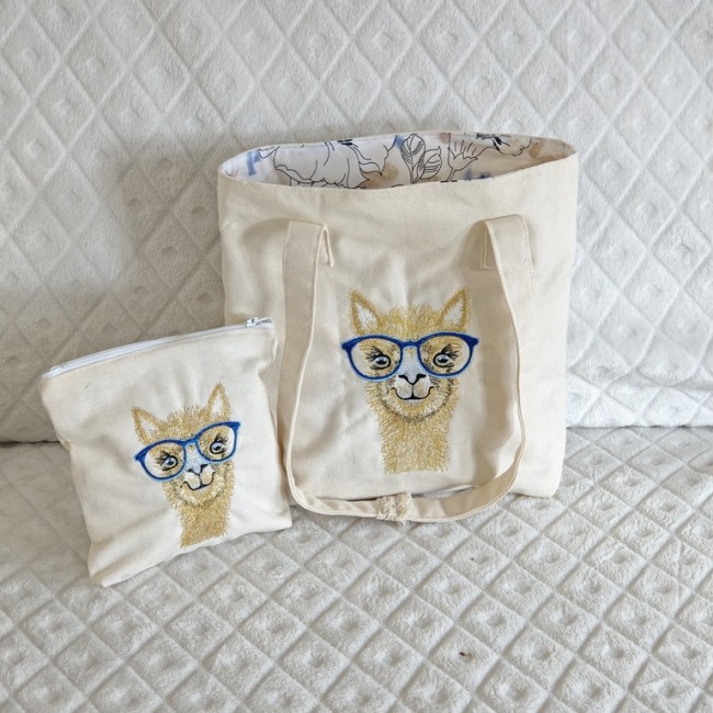 Ensemble sac et pochette en toile avec lama brodé – création artisanale originale Sac et pochette en toile beige avec broderie d’un lama à lunettes bleues, posés sur tissu matelassé.