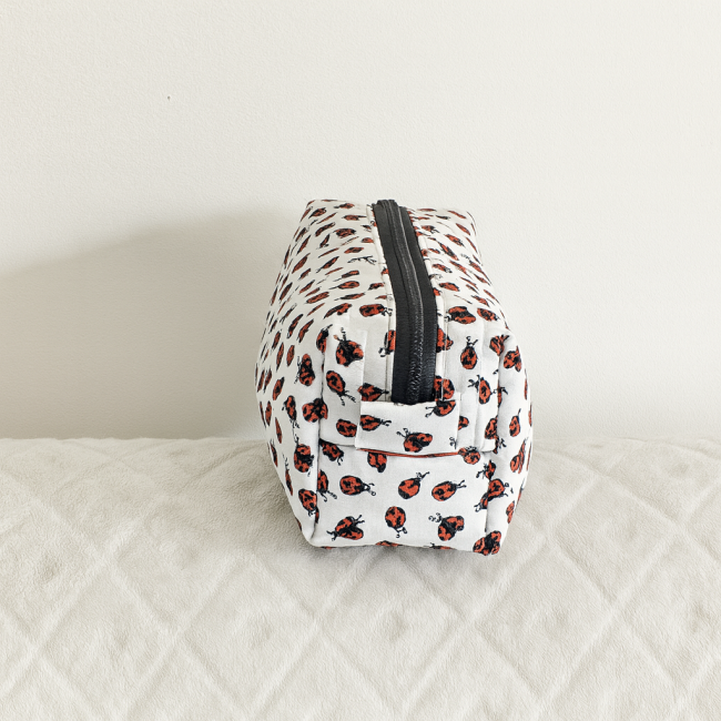Trousse rectangulaire blanche avec imprimé coccinelles rouges et noires, posée sur tissu matelassé.