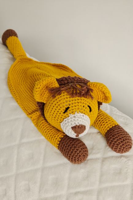 Lion en peluche crocheté à la main, jaune et brun, avec museau blanc, posé sur fond matelassé