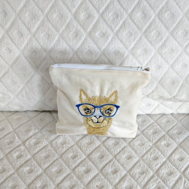 Pochette artisanale en toile avec broderie lama – accessoire ludique et pratique. Pochette en toile beige avec broderie d’un lama à lunettes bleues, posée sur tissu matelassé blanc.