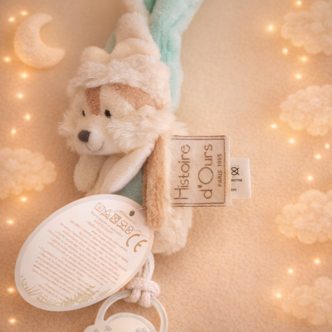 Attache‑sucette avec doudou blanc et bleu, clip sécurisé et étiquette ronde, présentée sur fond sable vanille avec lumière douce.