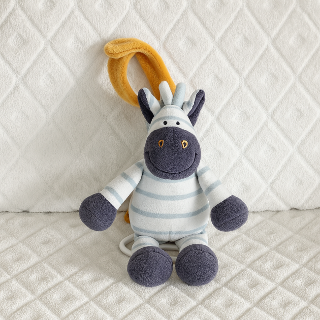 Boîte à musique Noukie’s – zèbre rayé bleu et blanc avec boucle jaune Peluche zèbre bleu et blanc Noukie’s assise sur couverture blanche, boucle jaune pour suspension