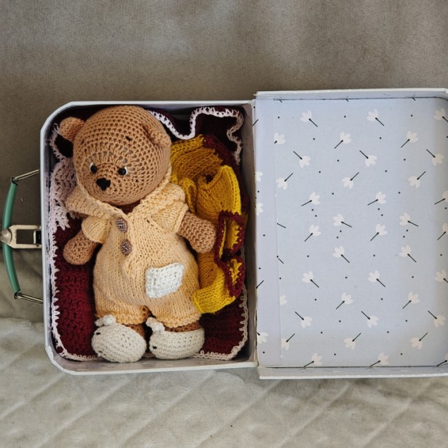 Ours au crochet dans sa valisette et accessoires artisanaux Ours au crochet installé dans une valisette ouverte, entouré de vêtements et couvertures faits main
