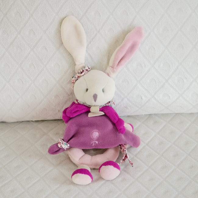 Lapin en peluche Doudou et Compagnie assis, tenue violette et rose, fond matelassé clair