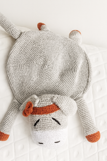Doudou crocheté en forme d’âne gris avec oreilles, pieds et nœud rouille, allongé sur une surface blanche.