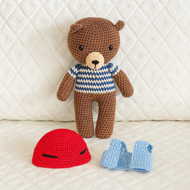 Ours en crochet fait main avec pull rayé bleu et blanc, casque rouge et gilet bleu posés devant