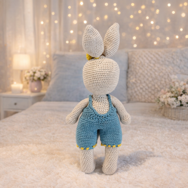 Peluche lapin crochetée en beige avec salopette bleue et nœud moutarde, posée sur un lit dans une chambre douce éclairée par des lumières chaudes.