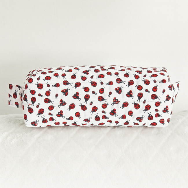 Trousse blanche à motif coccinelles avec anse latérale, posée sur surface matelassée.