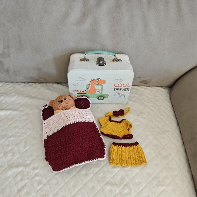 Ours au crochet et tenue complète présentés avec valisette Petit ours au crochet couché sous une couverture bordeaux et rose, accompagné d’une tenue moutarde et d’une valisette illustrée