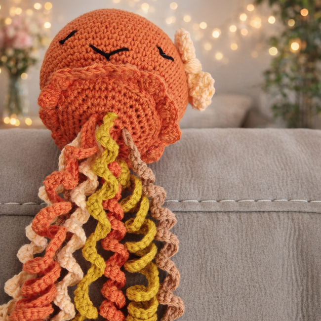 Pieuvre en crochet poétique – Bouille de Laine Pieuvre en crochet orange aux yeux fermés, ornée d’un noeud , avec tentacules bouclés multicolores et décor flou végétal et lumineux