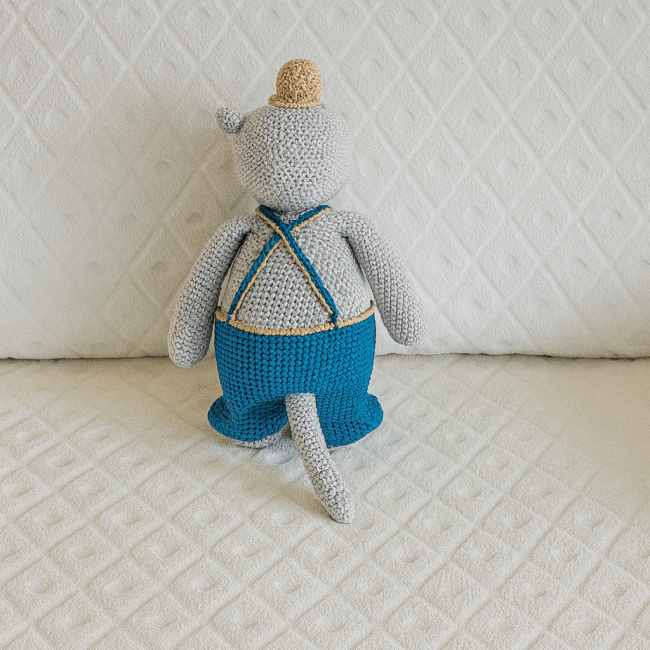 Doudou hippopotame crocheté en coton gris, salopette bleue et chapeau beige, vu de dos sur couverture blanche