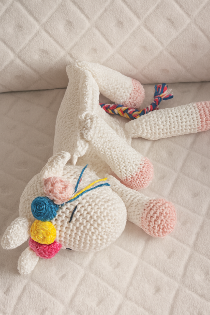 Doudou licorne en crochet avec crinière tressée et fleurs crochetées, posé sur tissu matelassé