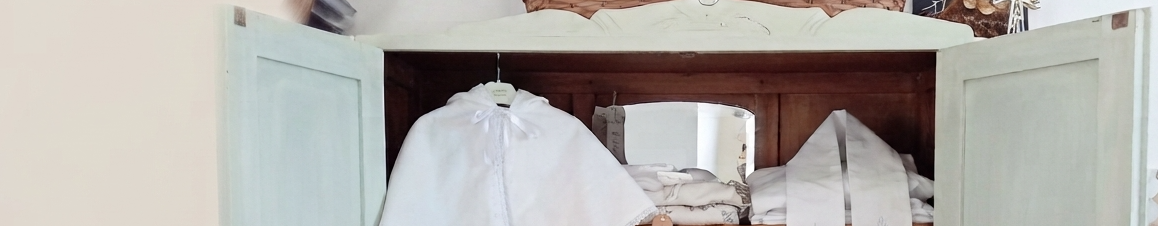 Armoire ouverte présentant des capes et écharpes blanches de baptême confectionnées à la main