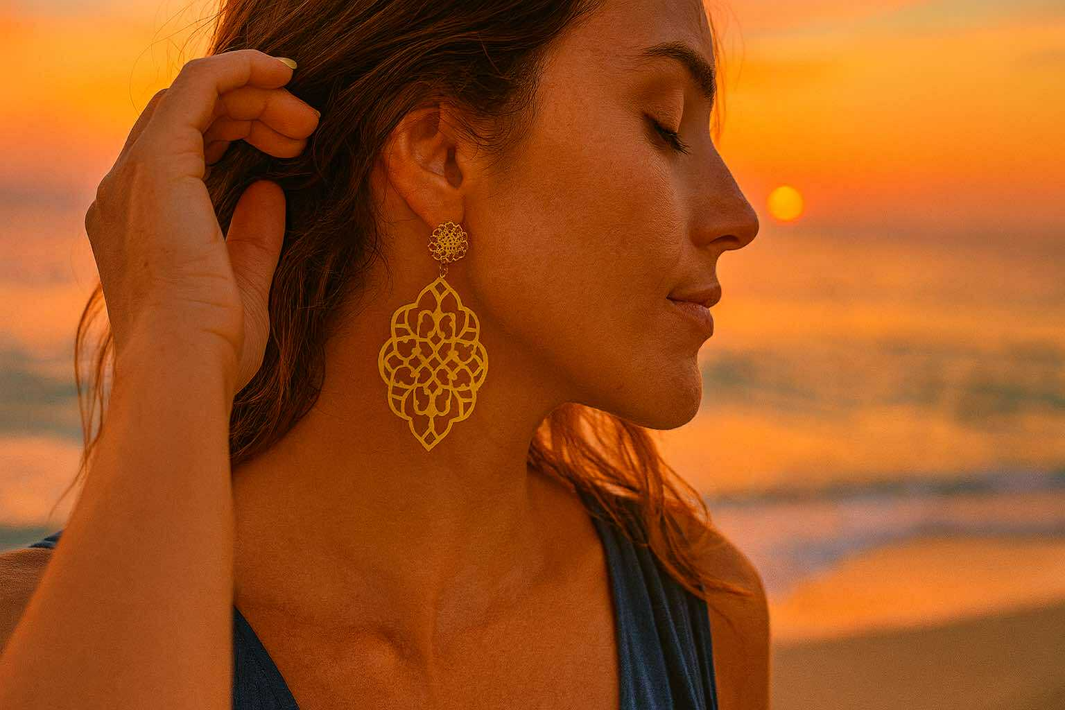 Femme portant de grandes boucles d’oreilles dorées ajourées au coucher du soleil – bijoux bohèmes et solaires Caprissa Créations