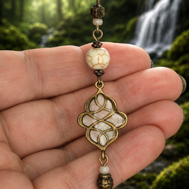 Détail du pendentif aux nœuds celtiques émaillés des boucles d’oreilles La Brume de Mimir. Laiton couleur bronze vieilli, émaillage nacré blanc artisanal et perles décoratives dans une atmosphère naturelle inspirée des forêts nordiques et des cascades anc