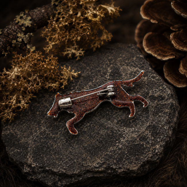 Vue arrière d’une broche artisanale représentant un loup viking inspiré de Sköll, avec attache métallique et émail cuivré scintillant, posée sur une pierre dans une ambiance nordique sombre.