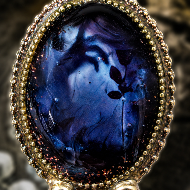 Gros plan sur le cabochon de la création artisanale Celle qui demeure, révélant ses nuances profondes bleu nuit, ses motifs mystérieux et son encadrement en laiton travaillé.