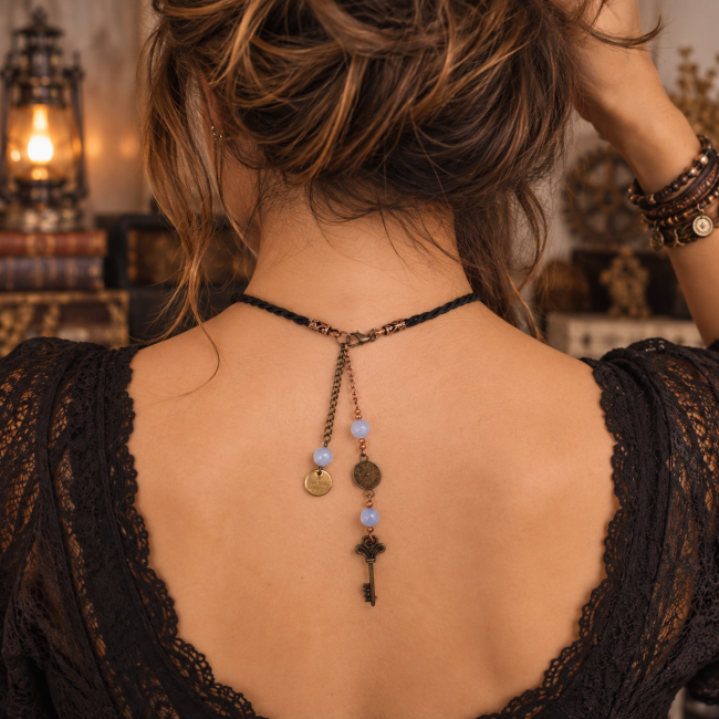 Vue portée au dos du collier Le Gardien des Archives, création artisanale de la collection Les Archives de Noctis. Le cordon tressé noir, les chaînes décoratives, les médaillons anciens, la clé et les perles de calcédoine bleue composent un bijou d’inspir