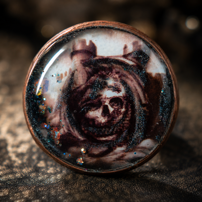 Cabochon émaillé La Crypte Oubliée – Crâne médiéval Gros plan du cabochon émaillé à la main de la bague La Crypte Oubliée représentant un crâne dans un paysage médiéval