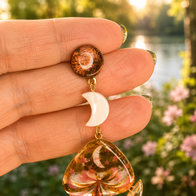 L’Éclat des Fées, des boucles d’oreilles artisanales inspirées de l’univers féerique, associant lune blanche, motifs floraux et émaillage aux reflets lumineux.