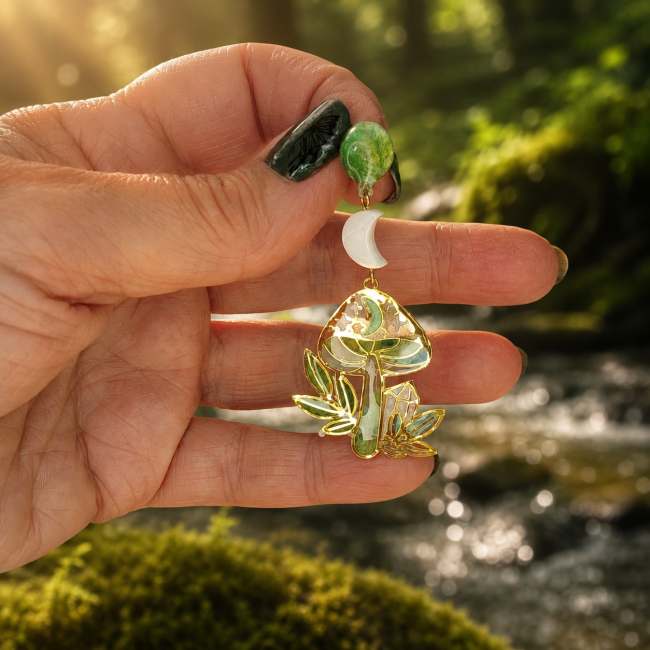 Détail artisanal — Nacre et émaillage vert des boucles Les Hindrès Boucle d’oreille Murmures des Sous-Bois présentée en main, champignon féerique et feuillage émaillés à la main dans un camaïeu de verts