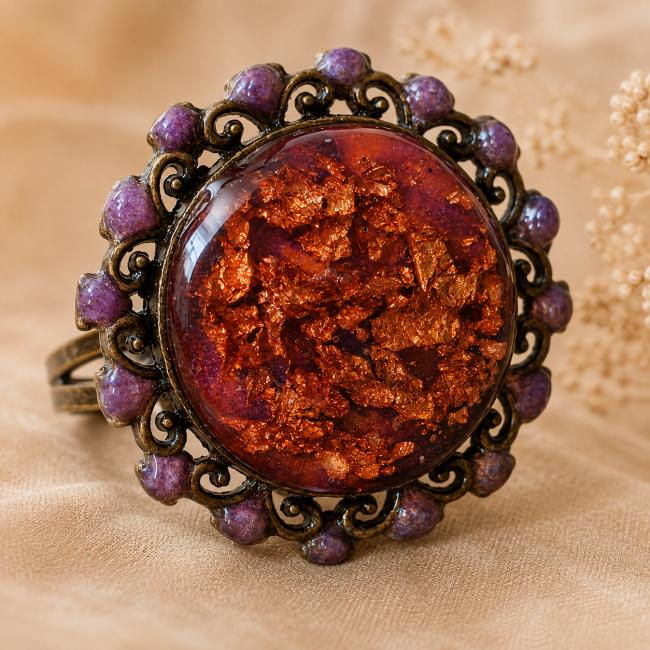 Zoom sur le grand cabochon travaillé et émaillé de la bague, mettant en valeur ses inclusions de feuilles de cuivre et son entourage délicatement émaillé violet.